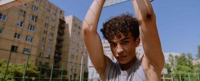 Teenage boy with curly hair in a gray tank top raising hands outdoors on a sunny day.