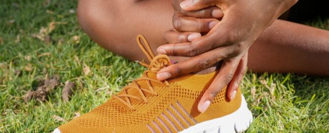 Close-up of athlete holding ankle with running shoe on grass, indicating pain or injury.