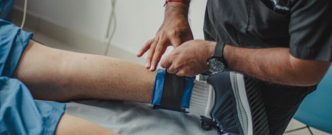 A physiotherapist adjusts a leg strap on a patient in a clinical setting.