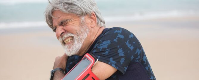 Elderly man grimacing from shoulder pain during seaside walk, showcasing health issue outdoors.